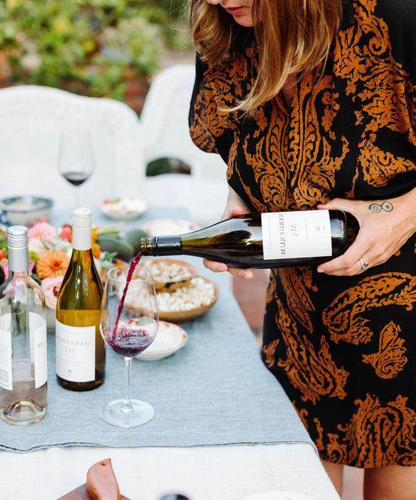 A woman pours Margerum Santa Barbara County M5 Rhone Blend red wine into a wine glass on a table full of appetizers in Santa Barbara